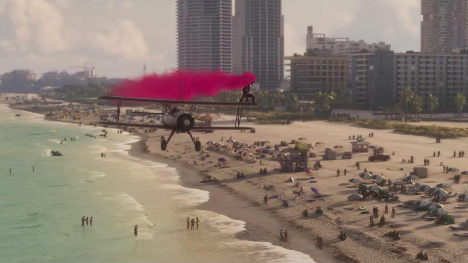 A red biplane trails pink smoke over a crowded beach, with the sea on one side and high-rises behind.