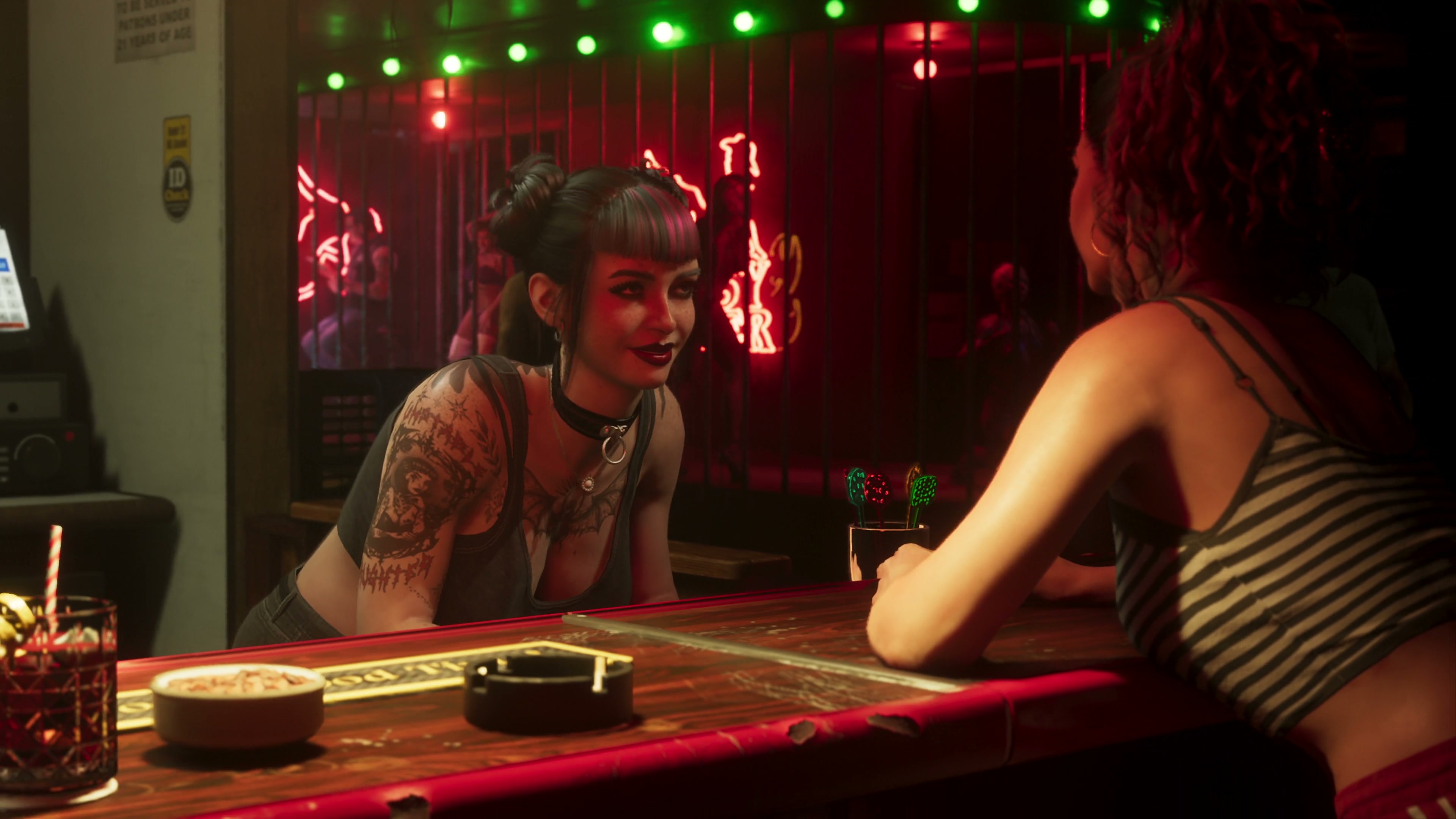Mujer de estilo goth con tatuajes se inclina sobre una barra en rojo y verde; frente a ella Lucía, de espaldas.