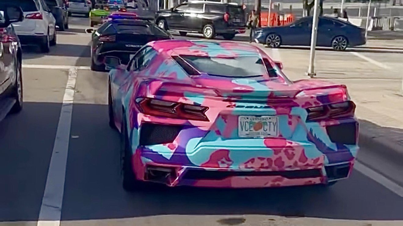 Corvette with a pink, blue and purple wrap and a "VCE CTY" plate, stopped at a downtown Miami light; ahead, a black Lamborghini with police lights