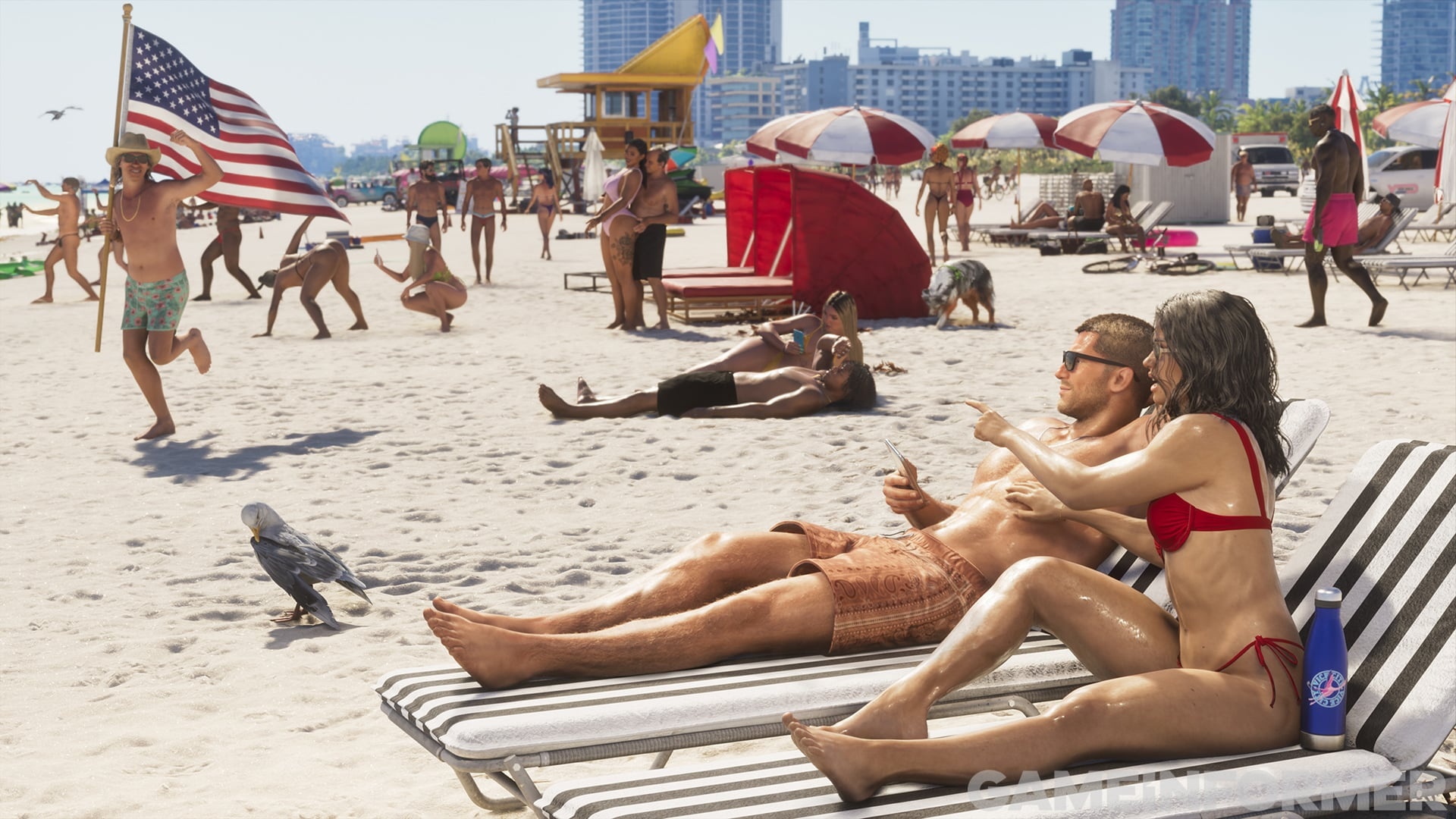 Crowded beach with a US flag, red umbrellas and a couple sunbathing on loungers in the foreground.