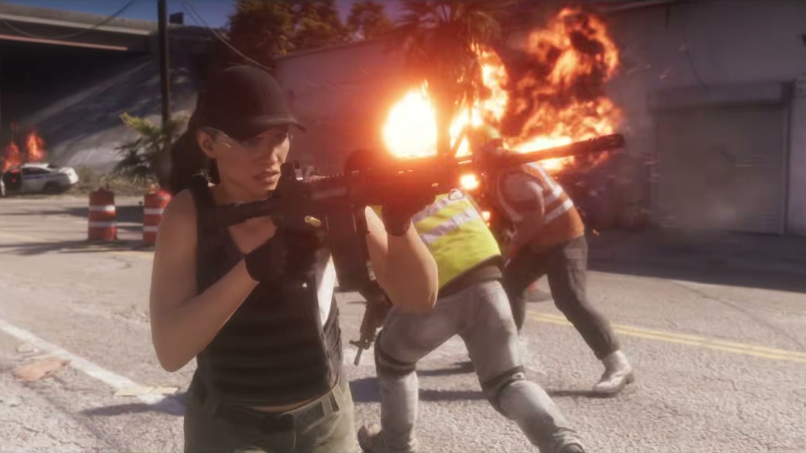 A woman in a cap and vest advances with an assault rifle down a street with fire and running workers.