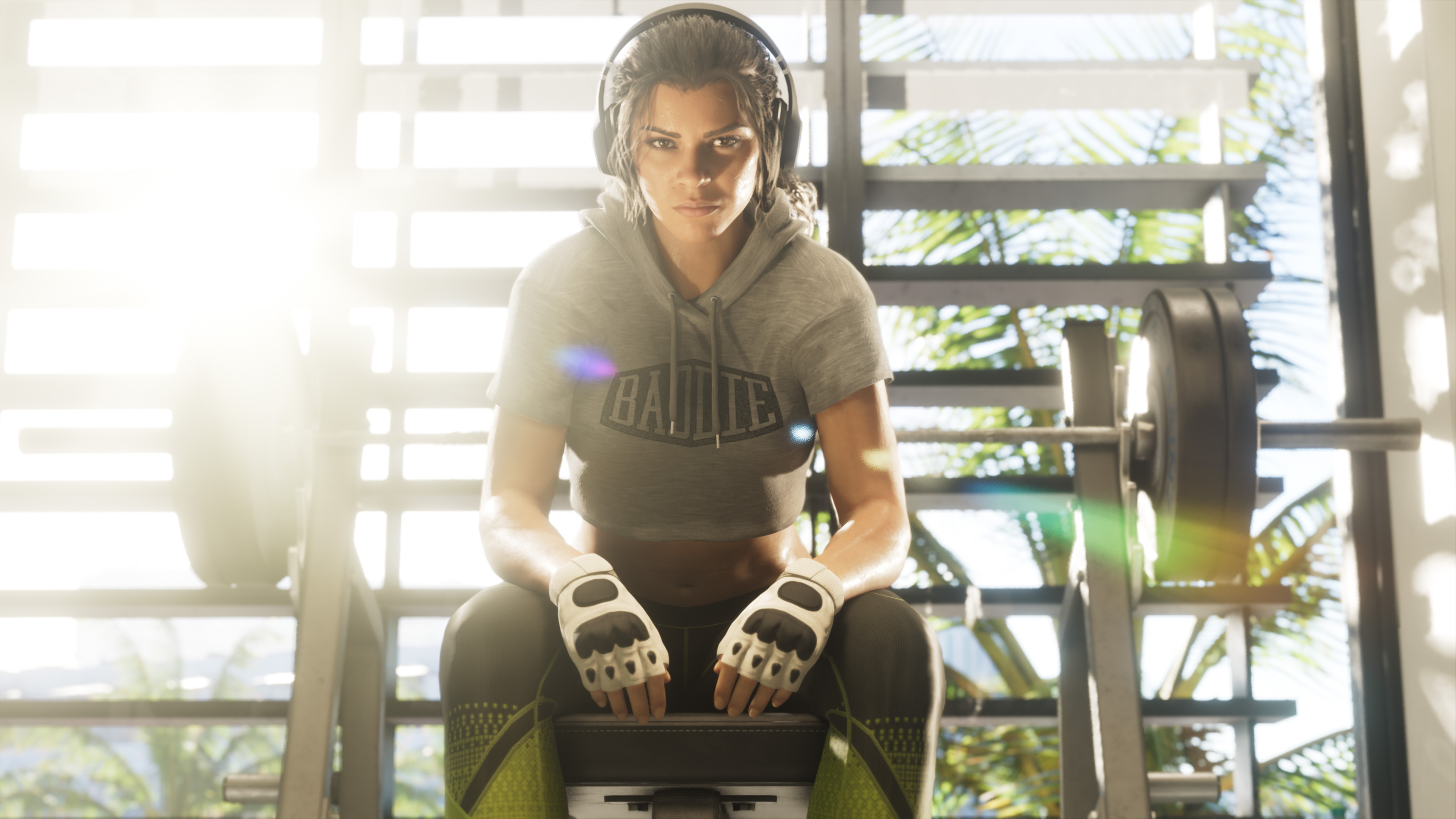 Woman with headphones, a Baddie hoodie and gym gloves sitting in front of a barbell; palm trees behind the glass.
