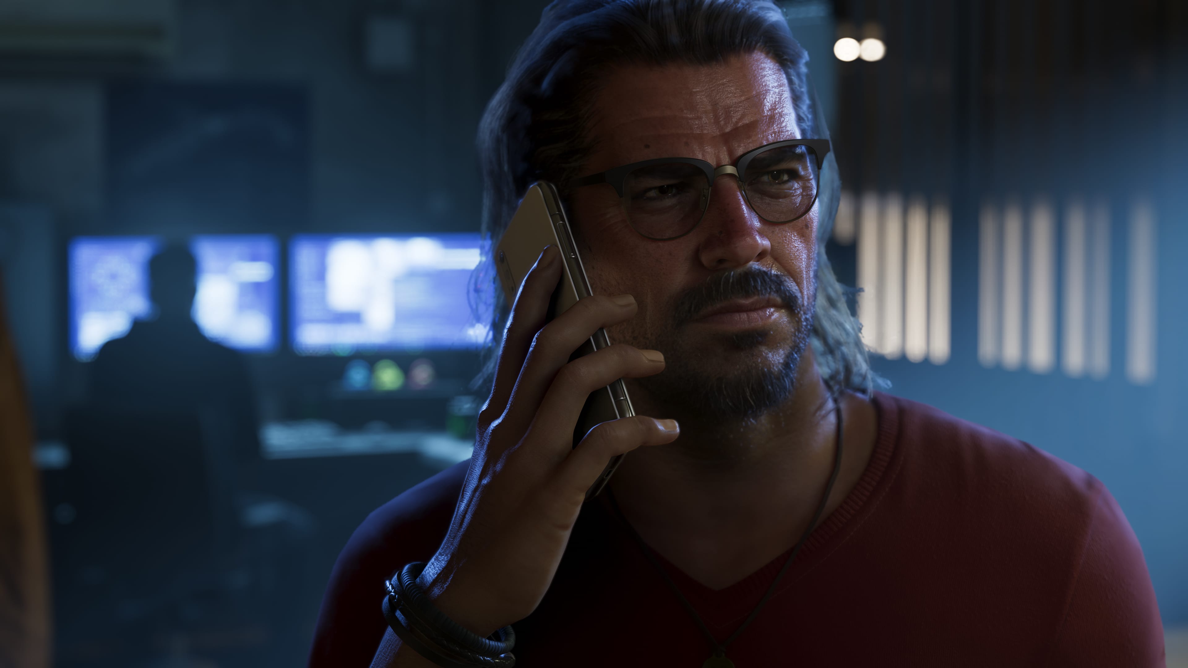 Man with glasses and a red sweater talking on a phone in a dark room, screens in the background.