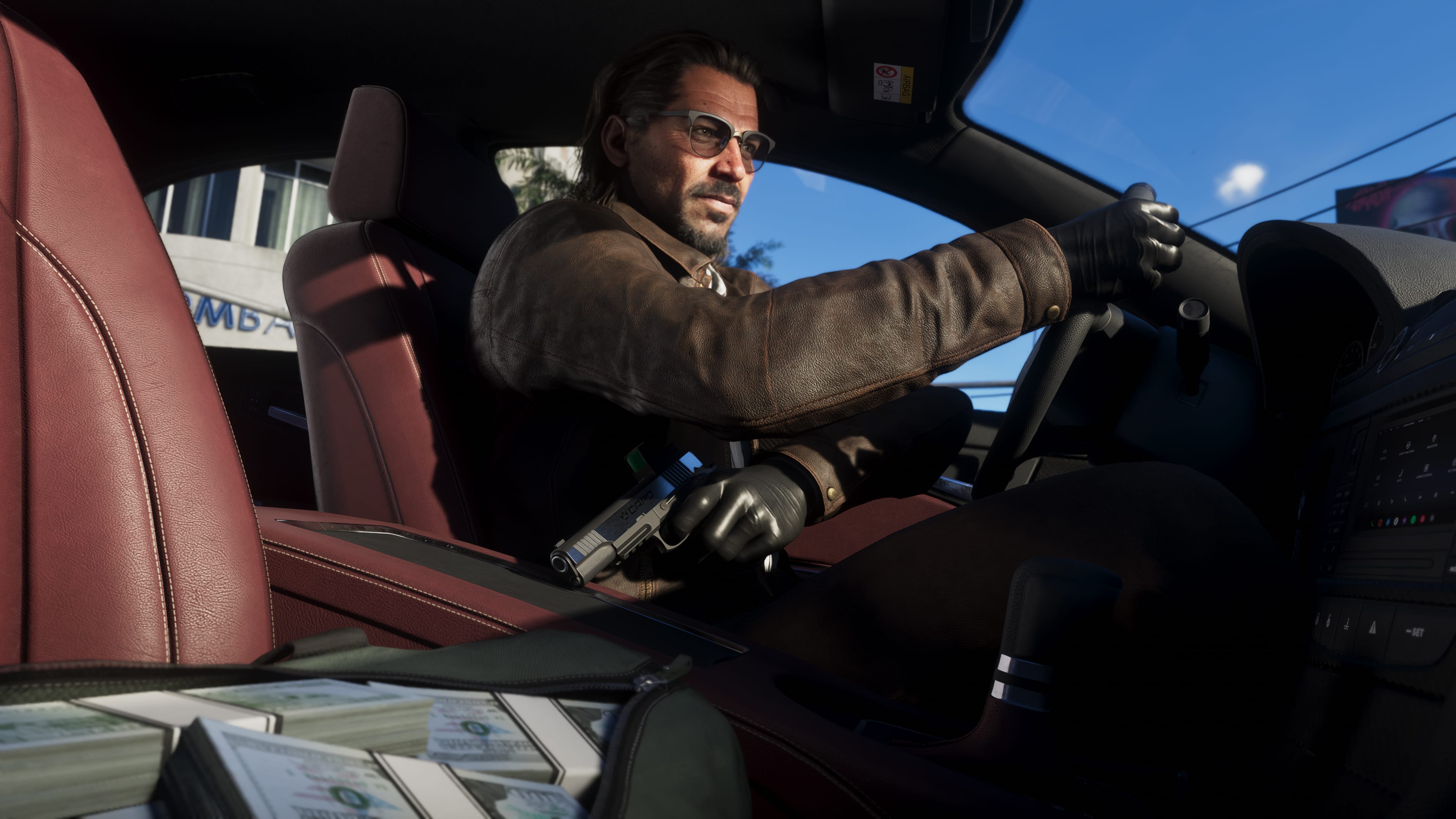 Man with glasses, a leather jacket and gloves driving a car with red seats; a pistol in one hand and stacks of cash in the foreground.
