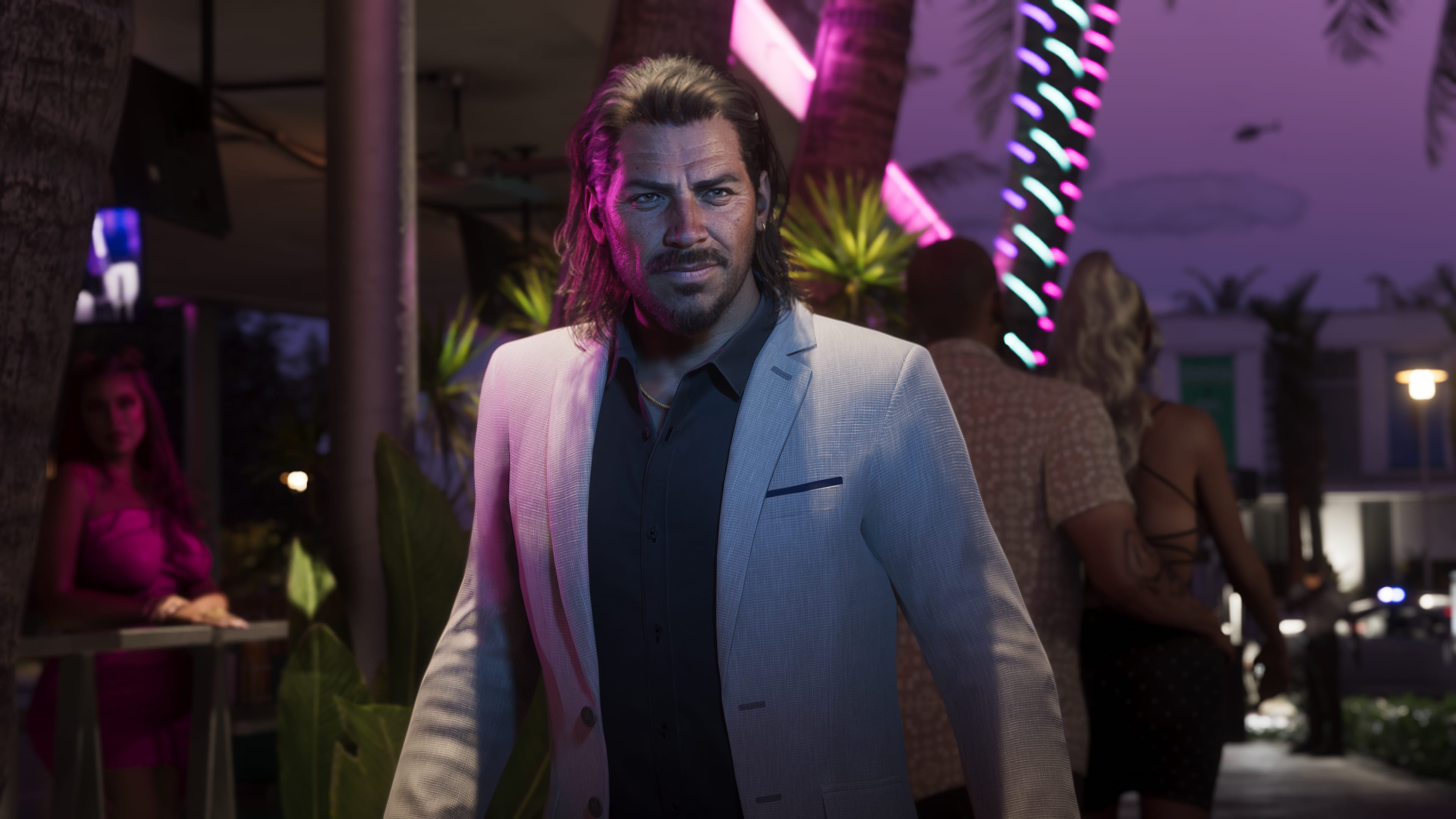 Man in a light jacket and dark shirt looking at the camera; pink neon lights and palm trees at night.