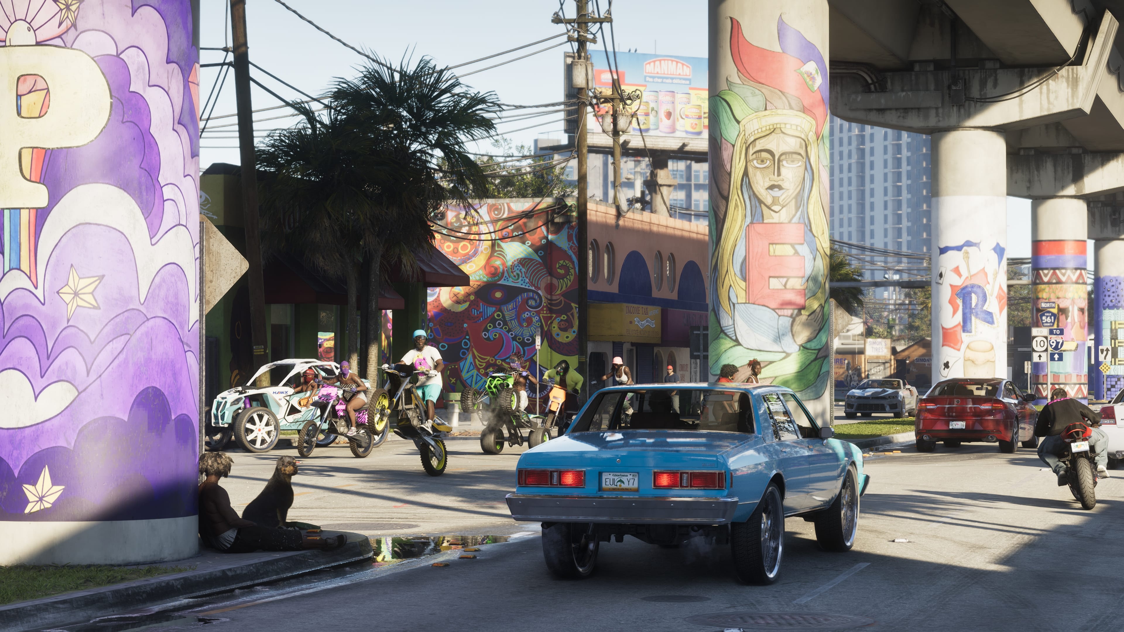 Classic blue car driving under a highway whose pillars are covered in murals, by day.