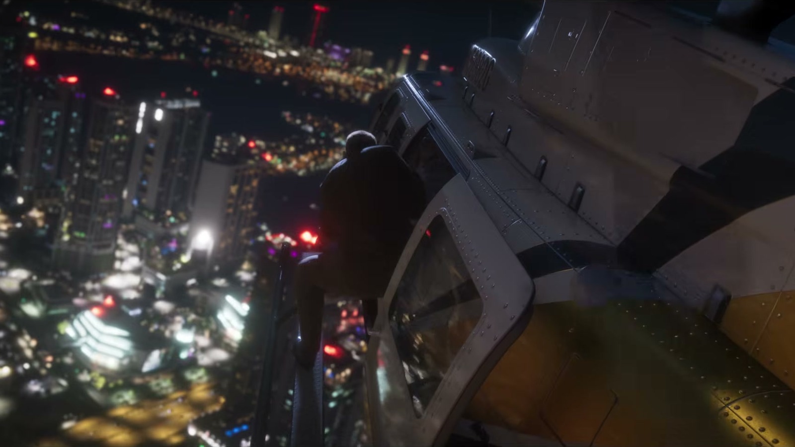 A figure clinging to the side of a white and yellow helicopter above the city at night.