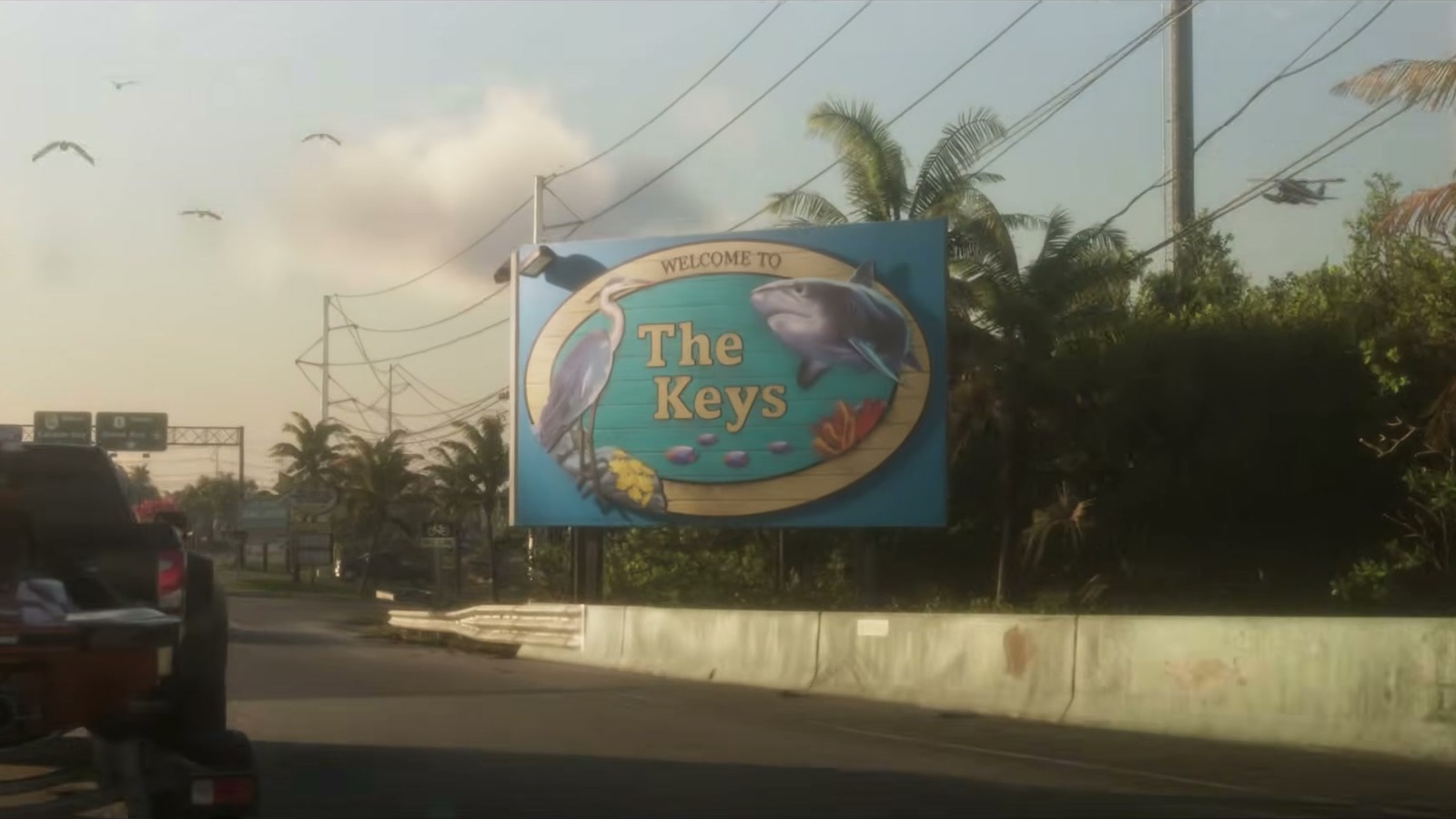 Welcome to The Keys billboard with a heron and a shark beside a palm-lined road.