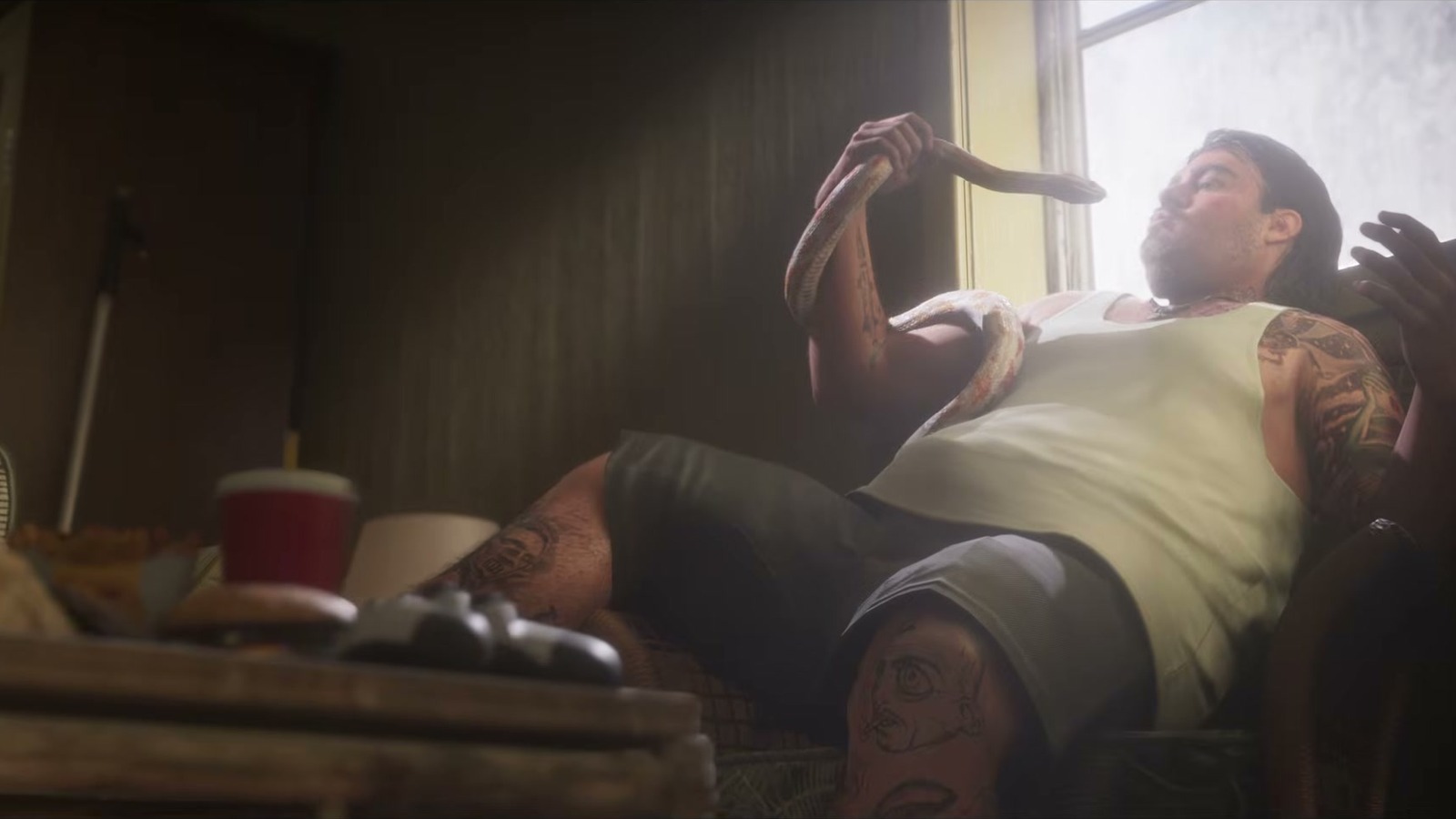A tattooed man in a tank top sits by a window with a snake wrapped around his arm.
