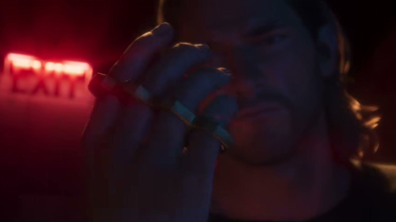 A bearded man puts on a brass-knuckle ring piece under red light, an EXIT sign behind him.